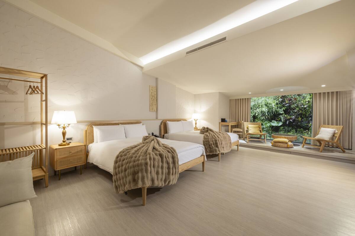 Habitación de hotel con dos camas, una zona de estar con una silla y una mesita, y vistas a zonas verdes al exterior.