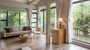A spacious living room with a sofa, a chair, a coffee table, and a side table with a lamp. Large windows offer a view of greenery outside. - Butterfly Valley Resort by Cosmos Otium (Wanrong)