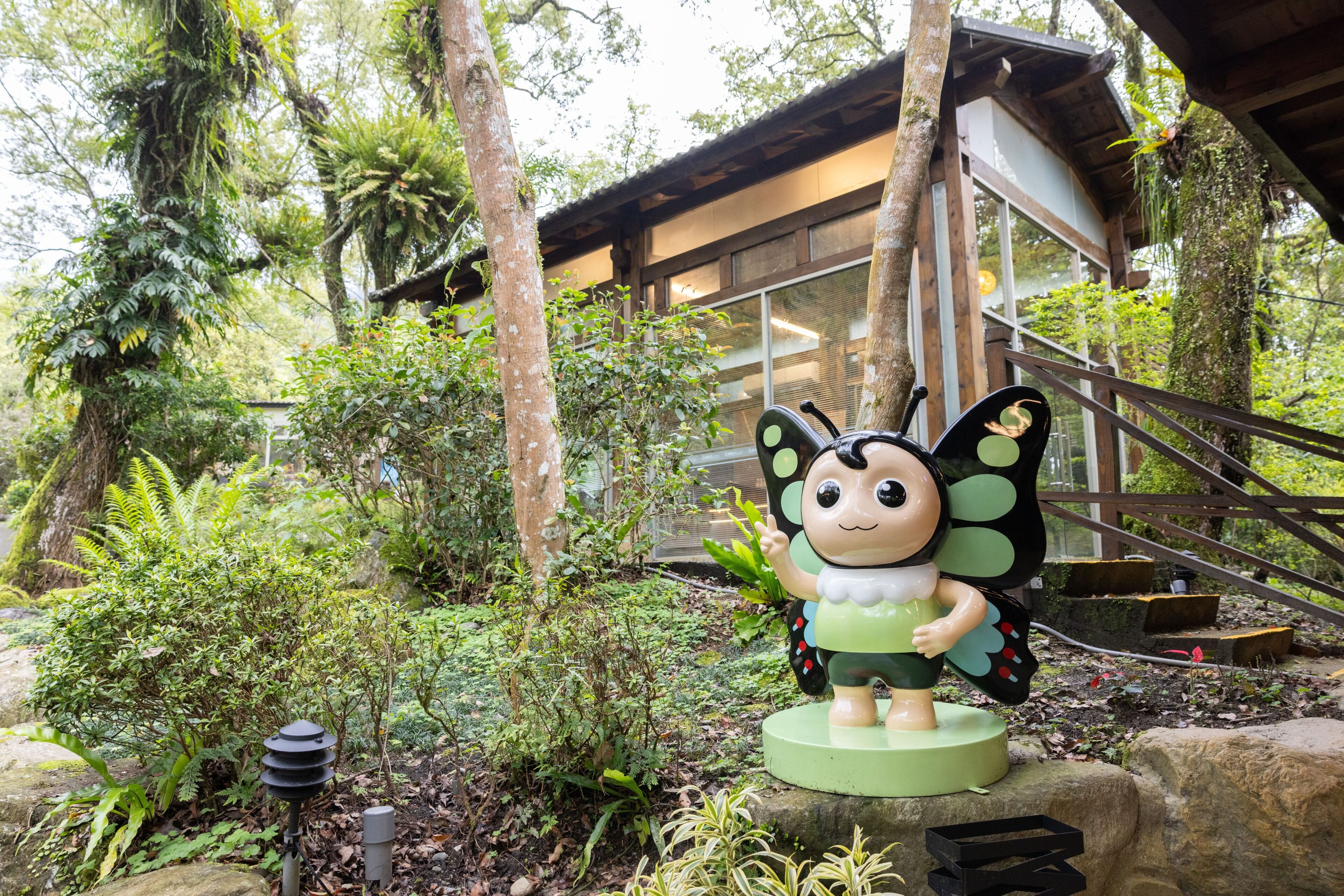 A statue of a character with a green dress and black wings, surrounded by lush greenery and a building with large windows.