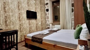 Hotellihuone, jossa on puinen sänky, työpöytä, seinälle asennettu televisio, ruukussa oleva kasvi ja ikkuna, jossa on verhot.