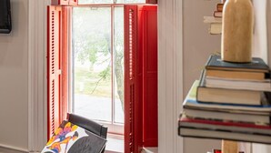 Una habitación con un gran ventanal con contraventanas rojas, una silla con un asiento colorido y una estantería con libros y objetos decorativos.