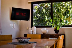 Une pièce avec un téléviseur affichant le logo « NETFLIX », une table à manger avec des chaises, un vase avec une plante et une fenêtre donnant sur un espace verdoyant.