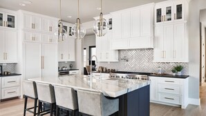 A modern kitchen with white cabinetry, a large island with bar stools, a herringbone tile backsplash, and pendant lighting.