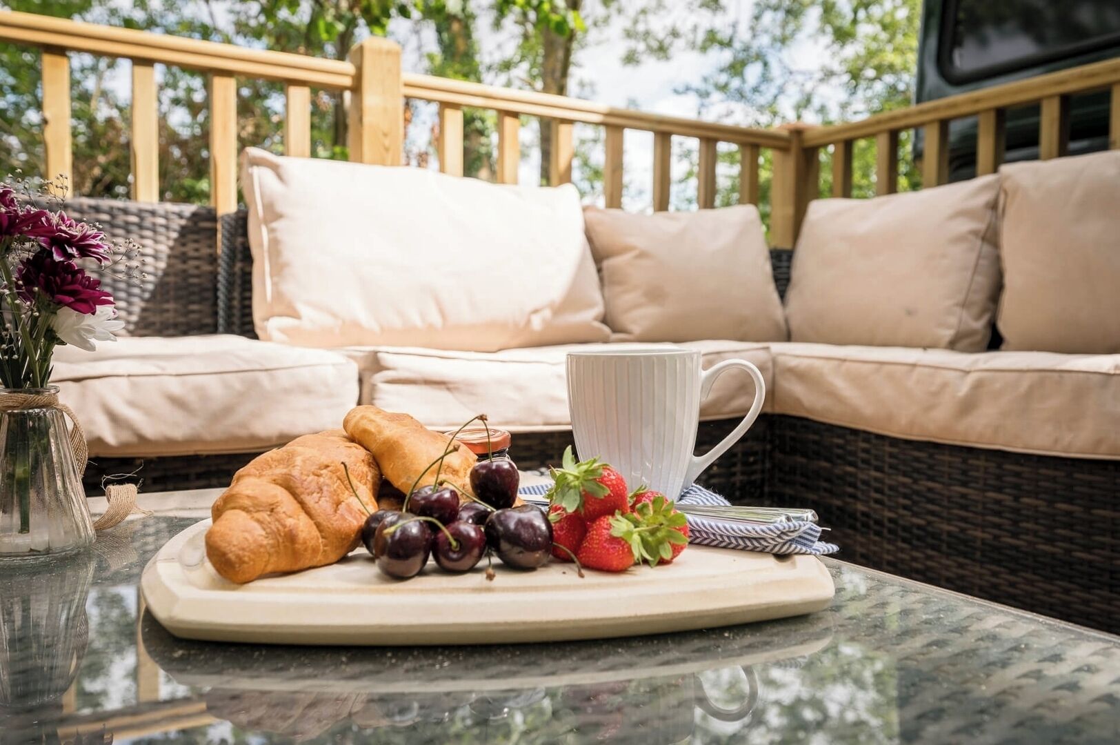 A breakfast tray with croissants, cherries, and strawberries on a glass table, with a wicker sofa and cushions in the background.