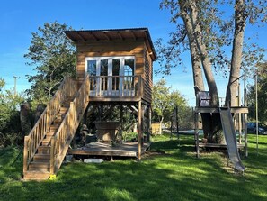 A wooden treehouse with a balcony and a slide, surrounded by trees and grass.