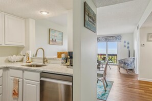 A modern kitchen with a stainless steel dishwasher, a sink, and a dining area with a view of the outdoors. - Ocean Racquet 8315 (St. Augustine)