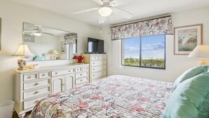 Una camera da letto con un letto, una cassettiera, una ventola a soffitto, una televisione, una finestra vista e opere d'arte.