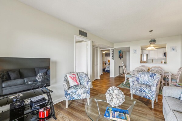 A living room with a TV, a sofa, and two patterned armchairs. There is a dining area with a table and chairs.