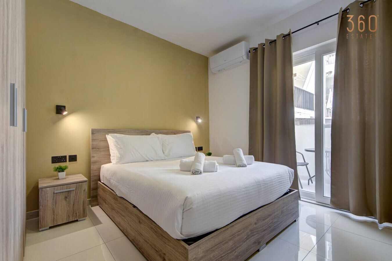Ein modernes Hotelzimmer mit einem hölzernen Bett, einem Nachttisch mit Pflanze und Blick durch große Fenster nach draußen.