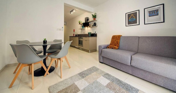 A modern living room with a grey sofa, a dining table with chairs, a kitchen in the background, and two framed pictures on the wall.