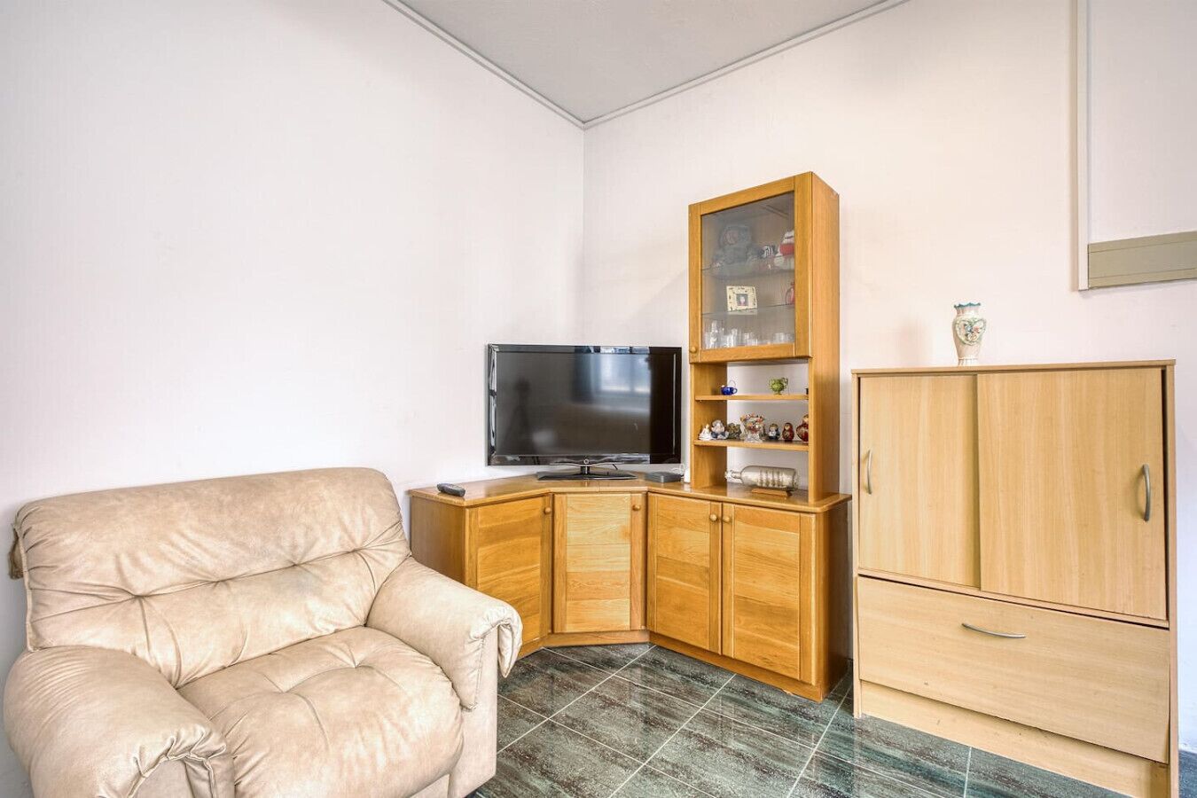 A living room with a beige sofa, a wooden cabinet with glass doors, a television, and a cabinet with a vase on top.