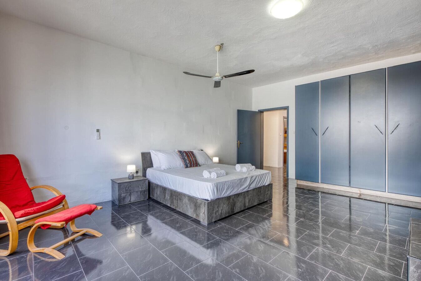 A bedroom with a bed, a red chair, a nightstand, a ceiling fan, and a wardrobe.