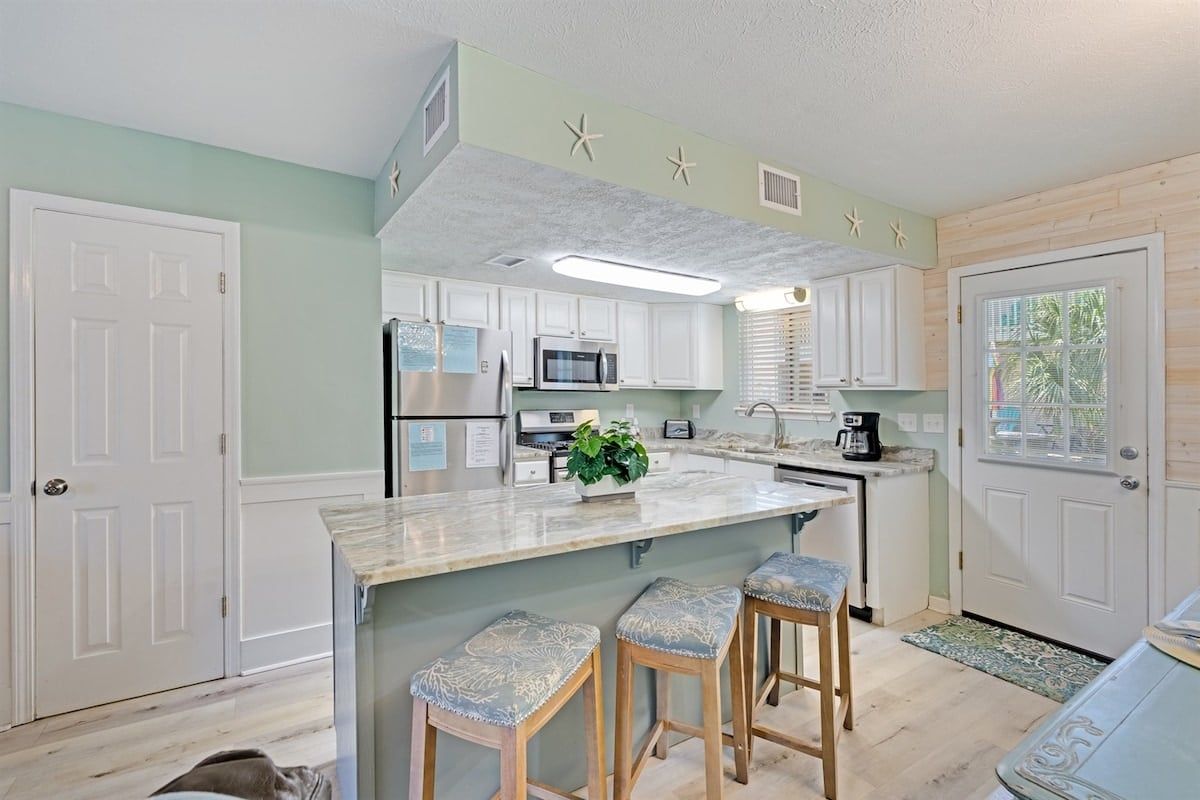 Una cocina con armarios blancos, una isla central con taburetes, un microondas y un refrigerador.