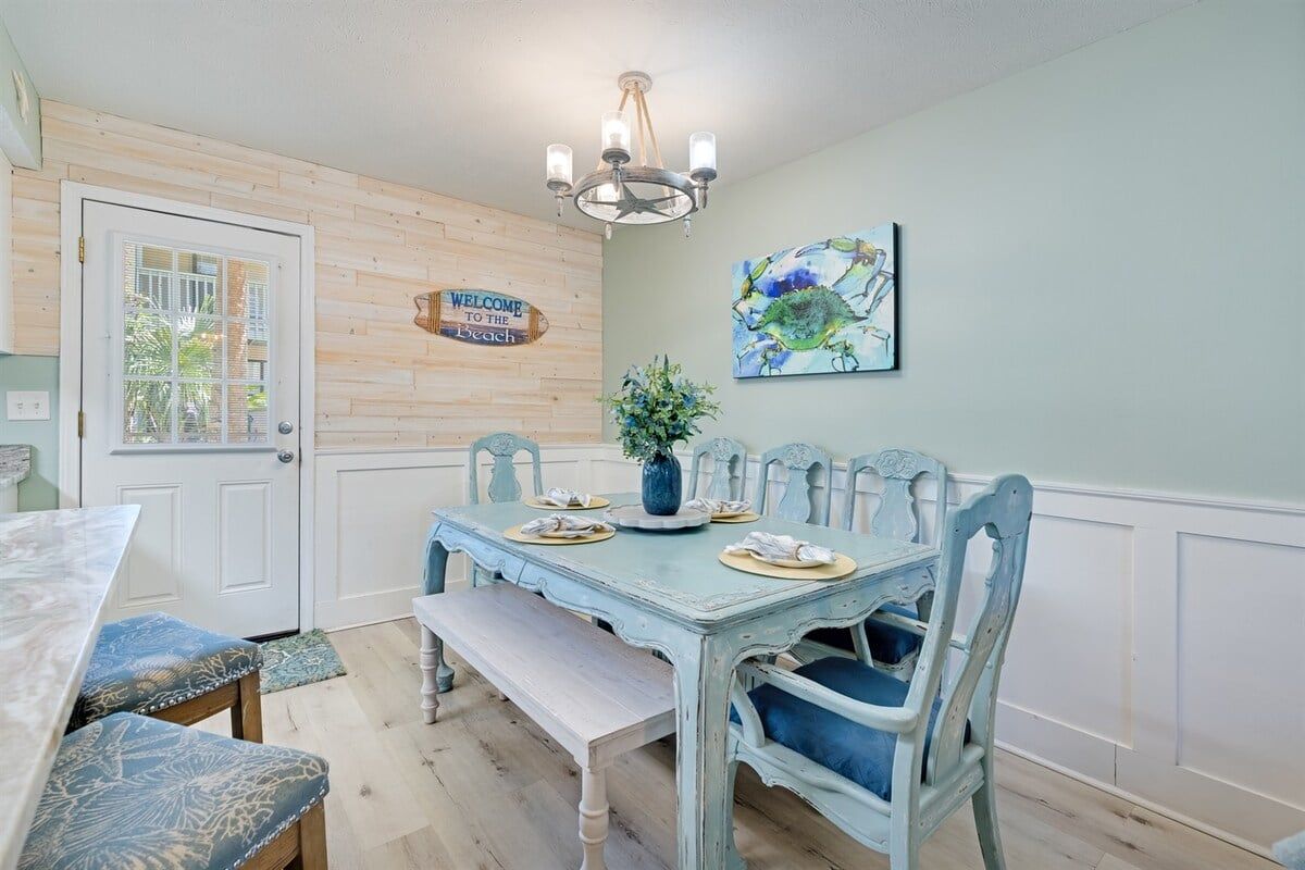 A dining area with a wooden door, a chandelier, a welcome sign, and a painting on the wall.