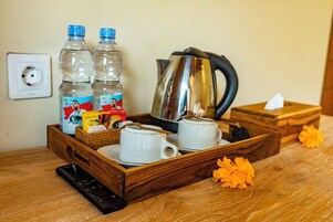 A wooden tray with two cups, two water bottles, a kettle, a basket with food items, a remote control, and a tissue box.