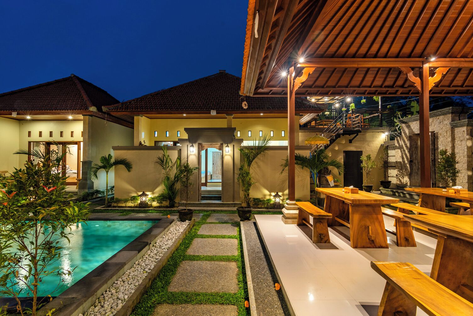 A poolside area with a wooden pergola, outdoor seating, and a staircase leading to a second floor.