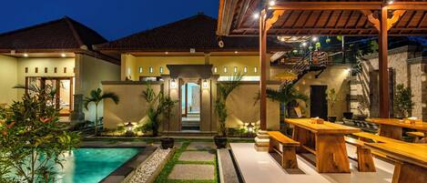 A poolside area with a wooden pergola, outdoor seating, and a staircase leading to a second floor.