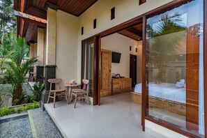 Un espace extérieur moderne, aménagé avec une table et des chaises en bois, un meuble en bois et un lit visible à travers une porte vitrée.