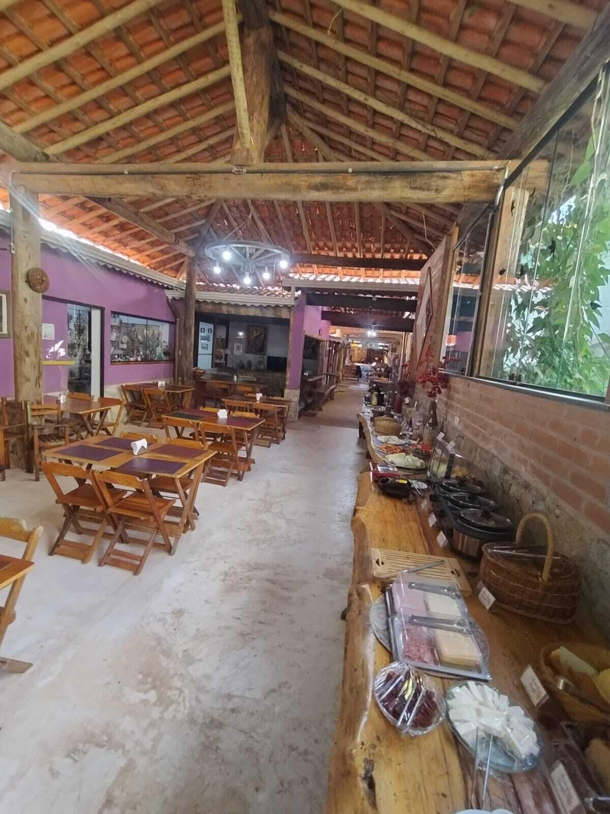 A rustic dining area with wooden tables and chairs, a brick wall with a window, and a ceiling with exposed beams.