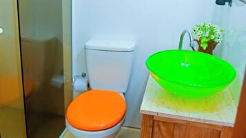 Une salle de bain avec une douche à parois de verre, des toilettes blanches avec un abattant orange et un meuble en bois avec un lavabo vert.