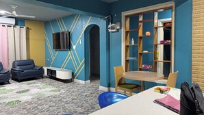 Een kamer met een blauw en geel geometrisch wandontwerp, een plafondventilator, een aan de muur bevestigde televisie, een kleine tafel met stoelen en een boekenkast met diverse voorwerpen.