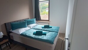 A bed with two pillows and a blanket, a bedside table with a lamp, and a window with a view of buildings. - Jessi`s Harz Apartment (Sankt Andreasberg)