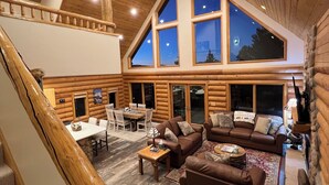 Un intérieur spacieux de chalet en bois, avec un plafond haut, de grandes fenêtres, un coin repas et un salon confortable.