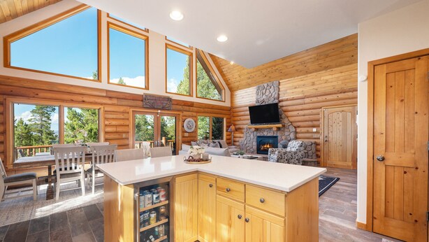 Un intérieur spacieux de chalet en bois, avec un grand îlot de cuisine, un coin repas et une cheminée en pierre.