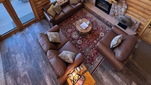 L’intérieur chaleureux d’un chalet en bois, avec une cheminée en pierre, des canapés en cuir et de grandes fenêtres donnant sur le paysage extérieur.