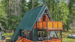 A two-story cabin with a green metal roof, wooden deck, and a small attached garage. - A-Frame @ Entrance to Rainier w/ Hot Tub & WiFi (Ashford)