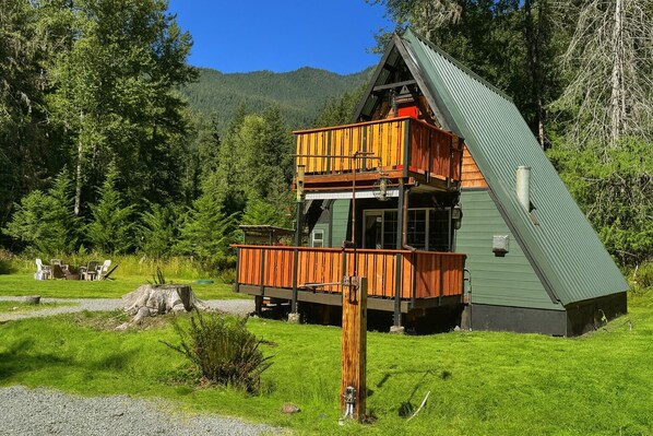A cabin with a green exterior and a wooden deck, surrounded by a forested area.