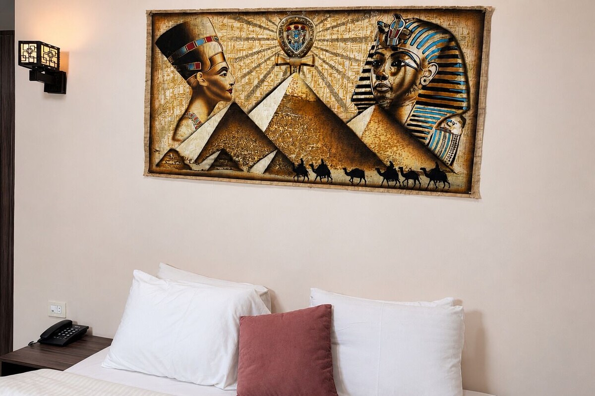 A hotel room with a bed, pillows, a nightstand with a phone, and a wall-mounted artwork of Egyptian figures.