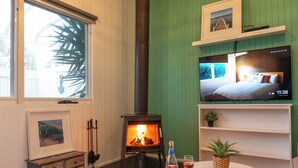 Ett mysigt rum med en vedeldad kamin, en tv som visar ett sovrum, ett litet bord med en flaska och ett glas samt en inramad tavla på en byrå.
