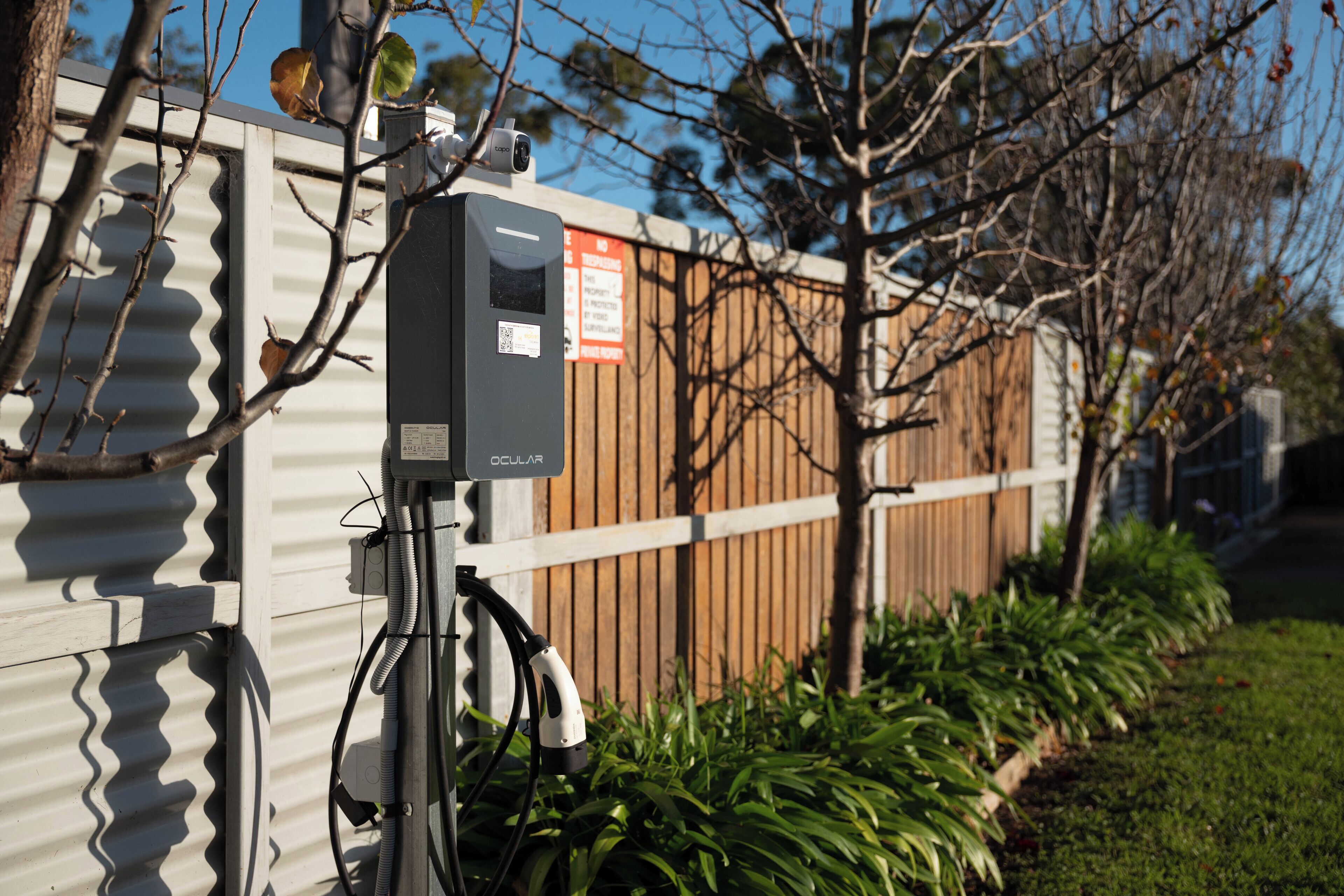A security camera mounted on a metal fence with a wooden panel, connected to a power box, and surrounded by greenery.