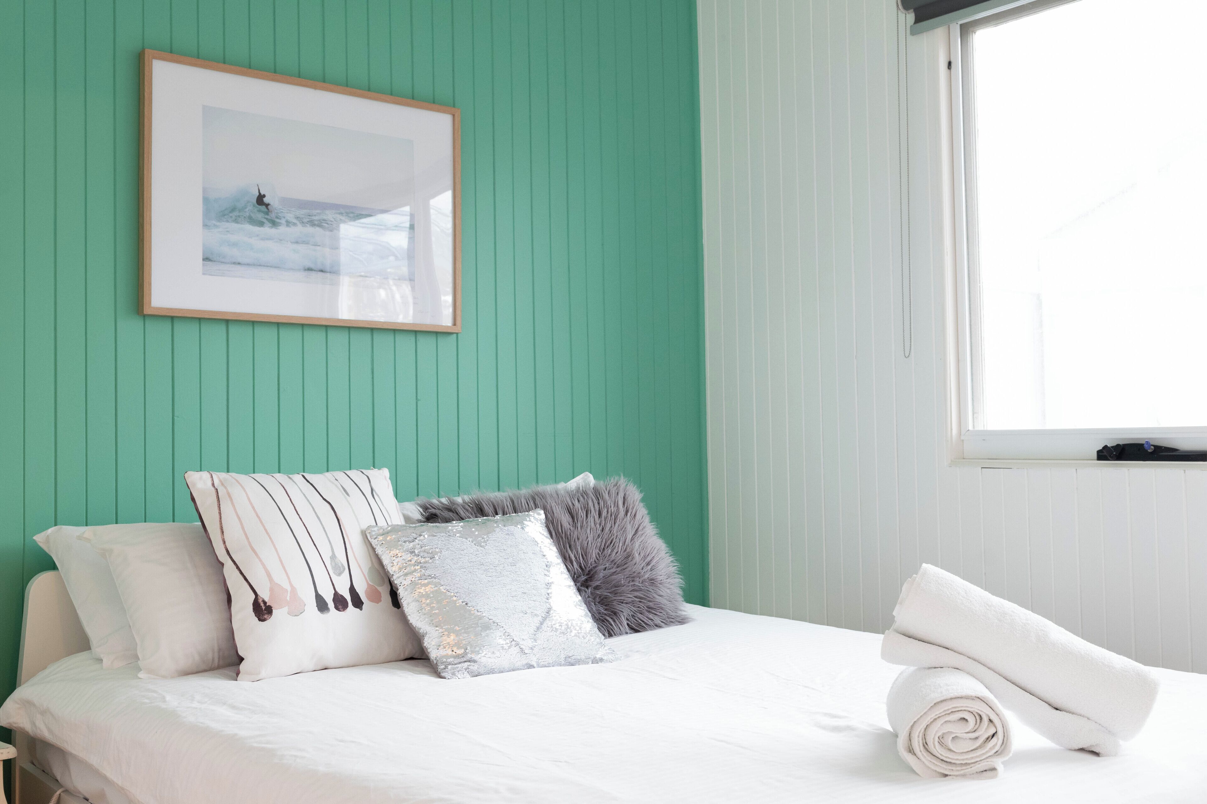 A neatly made bed with white linens, a framed picture on the wall, and a window with blinds.