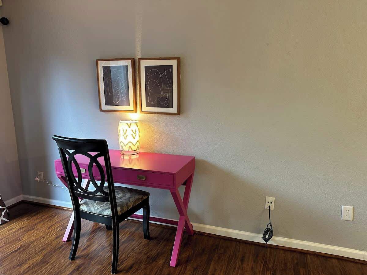 A room with a pink desk, a black chair, two framed abstract artworks on the wall, and a lamp on the desk.