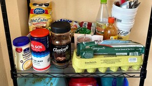 A kitchen shelf with various food items and kitchenware, including a jar of salt, a bottle of hot cakes, a can of cream substitute, a container of Classica cafe, a package of spaghetti, and several dishes and utensils.