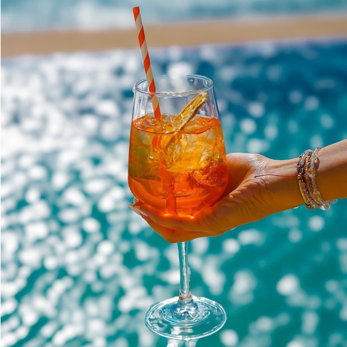 Eine Hand hält ein Glas mit einem Strohhalm, gefüllt mit einem Getränk mit Eiswürfeln und einer Zitronenscheibe, vor verschwommenem Hintergrund von Wasser.
