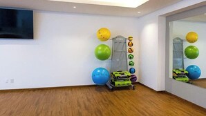 A room with a wooden floor, a television mounted on the wall, and exercise balls and equipment on a rack. - Encantame|2BR|Rooftop Pool|LazyRiver|Pickleball (Playa Encanto)