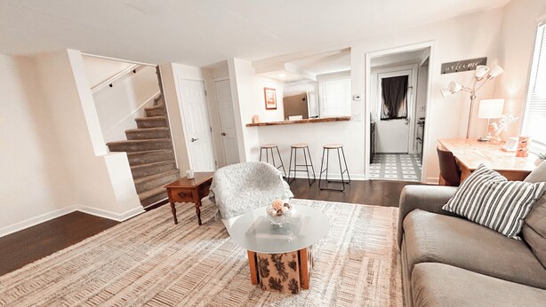 Un vaste espace de vie comprenant un canapé, une table basse et un tapis à motifs. Un escalier mène à l’étage supérieur, et on aperçoit un coin cuisine au fond.