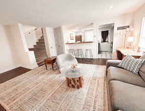 A spacious living area with a sofa, a coffee table, and a patterned rug. A staircase leads to an upper floor, and a kitchen area is visible in the background. - Private and Cozy 2 Bedroom House with Amenities! (Union Township)