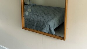 Un miroir avec un cadre en bois reflète un lit recouvert d’une couvre-lit à motifs et un mur orné d’un petit tableau encadré.