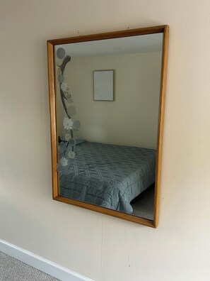Un espejo con marco de madera refleja una cama con edredón estampado y una pared con un pequeño cuadro enmarcado.