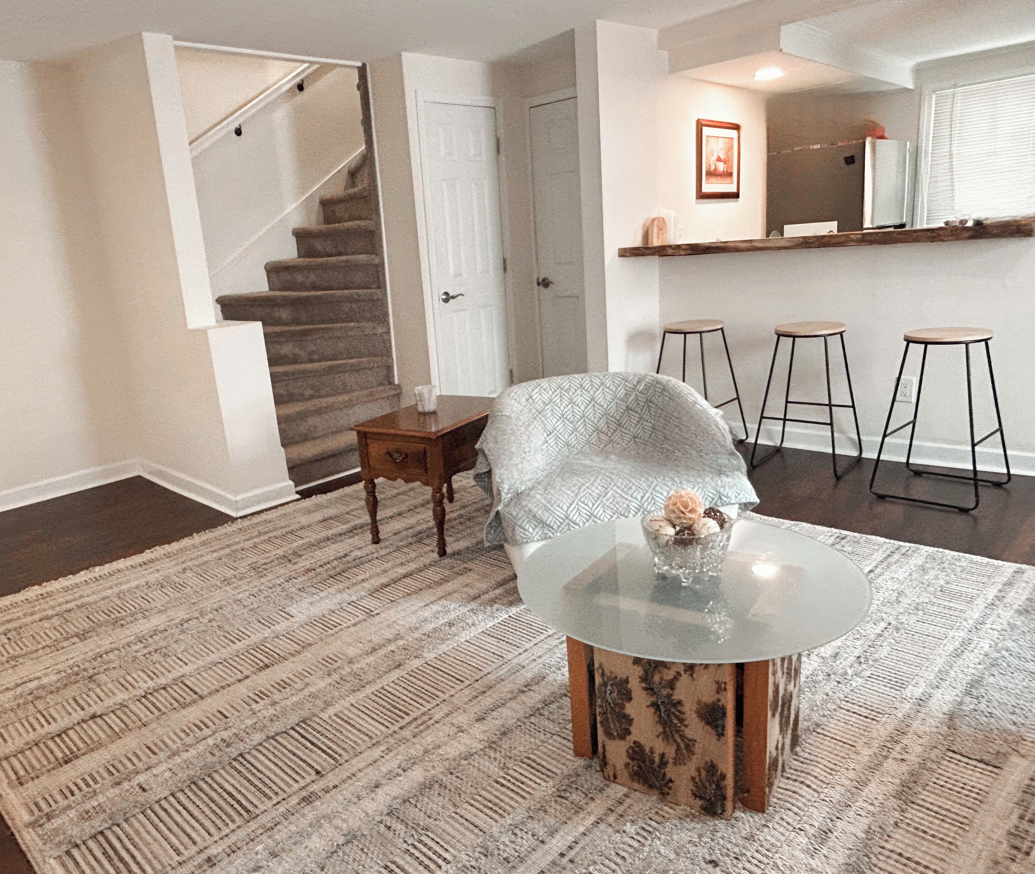 Un salón con una escalera, una mesa de centro de vidrio y una alfombra estampada.