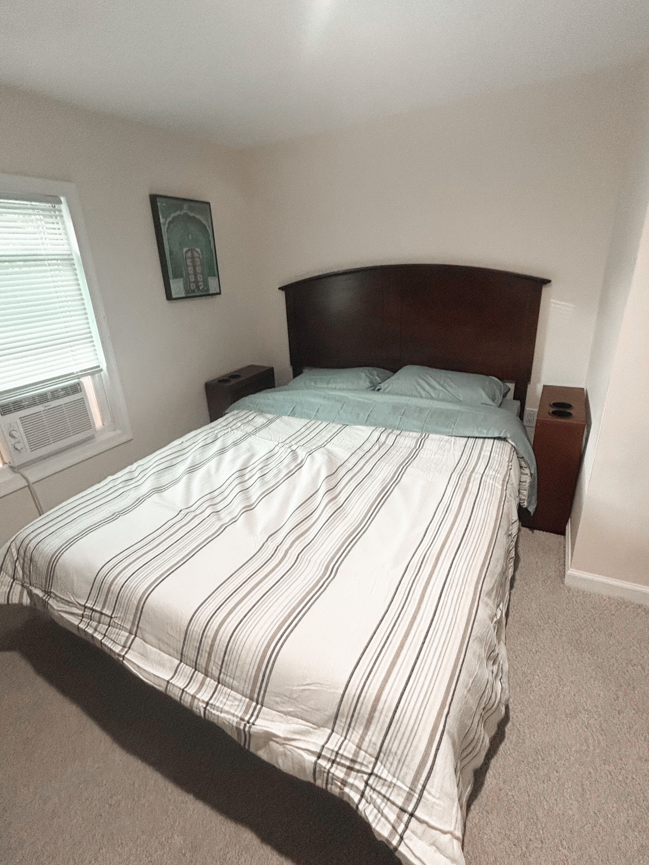 Quarto com cama, mesa de cabeceira, janela com persianas e um quadro na parede.