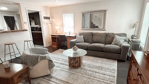 Un salon avec un canapé, une table basse et un tapis. On aperçoit un coin cuisine au fond de la pièce.