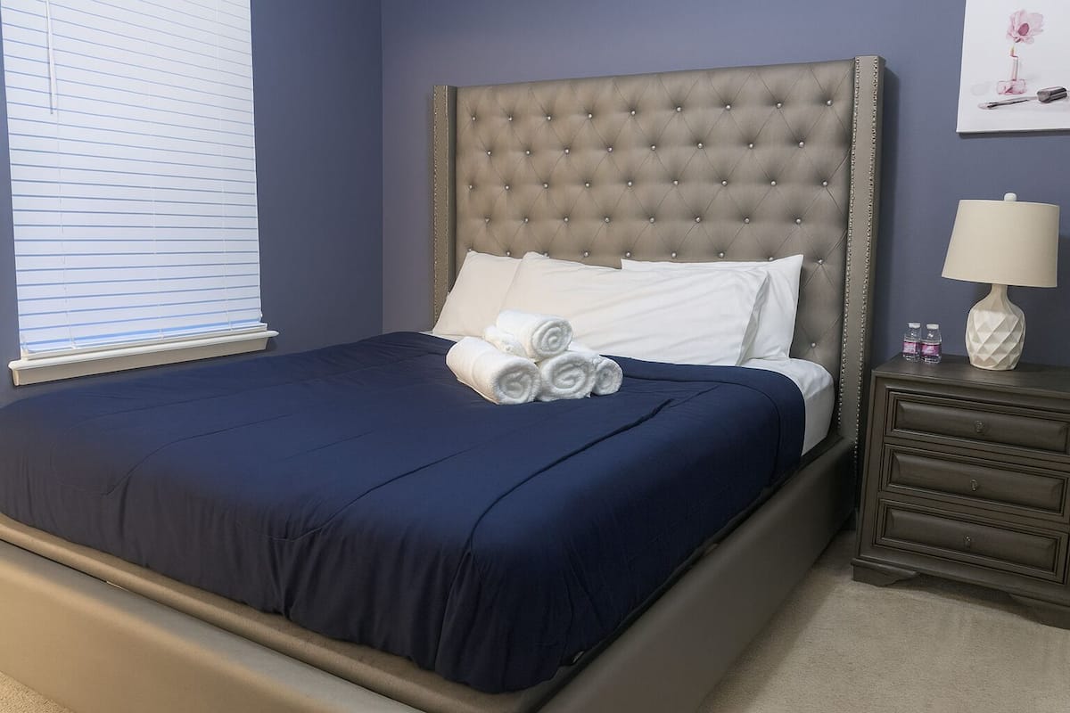 A neatly made bed with a dark blue bedspread and white pillows, a tufted headboard, and a nightstand with a lamp and small bottles.
