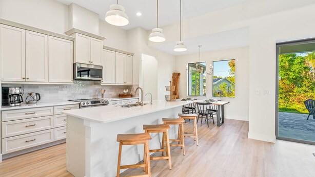 A modern kitchen with white cabinetry, a central island with bar stools, stainless steel appliances, and a large window offering a view of the outdoors. - New 3BR , Near Myakka River, Boca Grande + Beach (Port Charlotte)