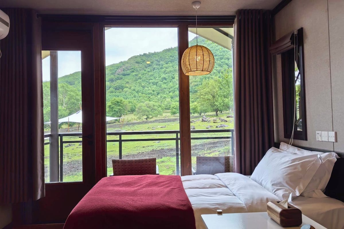 A hotel room with a bed, a small table, a hanging light fixture, and a view of a green landscape through large windows.