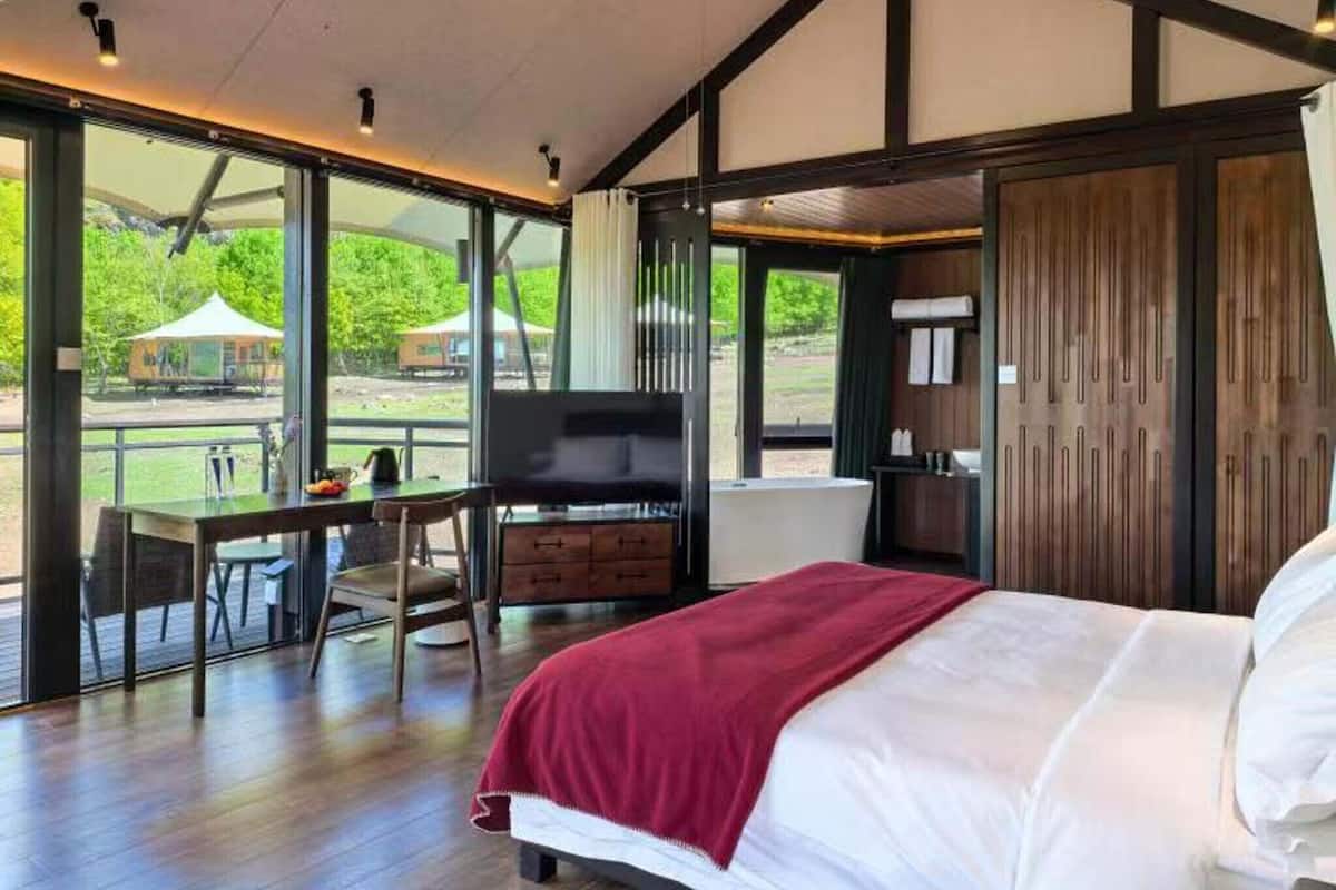 A bedroom with a bed, a desk, and a chair, large windows, and a view of the outdoors.
